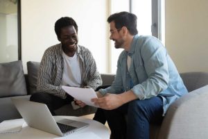 two men sit together reviewing documents on laptop and paper and defining what is financial coaching