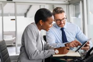 a woman sits and with a man who is showing something to her on tablet while she writes on a notepad during their financial coaching services session