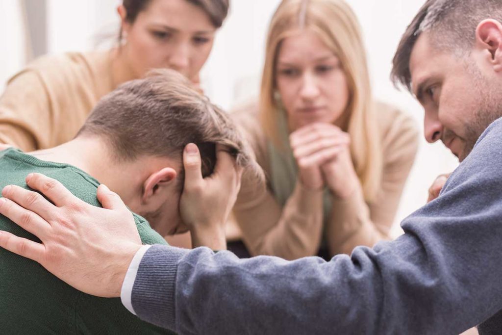 a man holds his hands to his head is upset while other people around him participate in alcohol interventions attempt to console the man