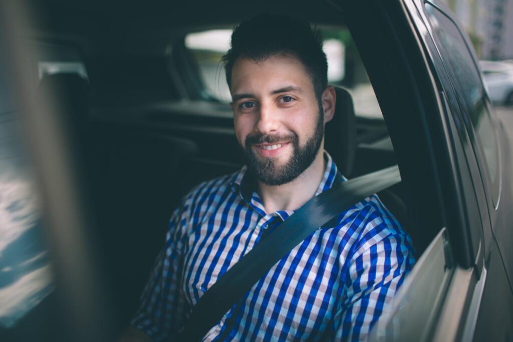 a man smiles while sitting in a car with seatbelt on and utilizing sober transport services