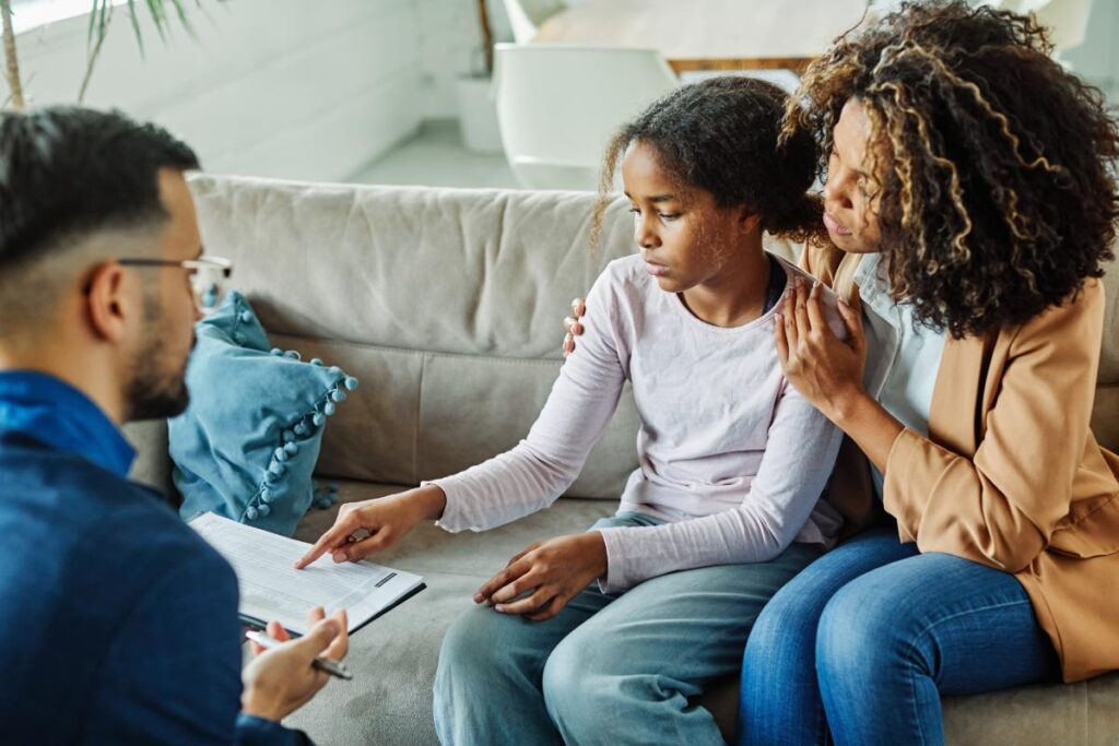 a woman sits on the couch with her young daughter who is pointing to the paper on a therapist's clipboard who is discussing interventions for family therapy