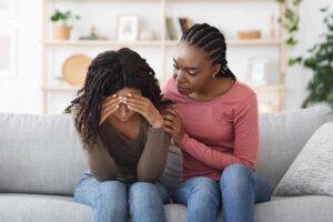 a woman sits on a couch next to another woman who has her head held down appearing distraught while listening to her friend discuss the benefits of early interventions