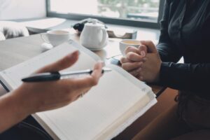 a person holds a pen while notebook sits on a desk while they explain benefits of recovery coaching to another person