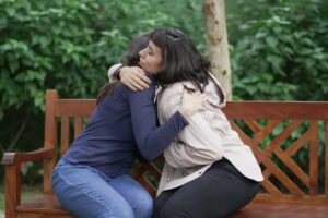 two women sit on a park bench outdoors and embrace after learning more about coping with your loved one in addiction treatment