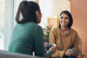 a woman sits in a chair holding a coffee cup while talking to a woman sitting across from her and asks who pays for sober living homes