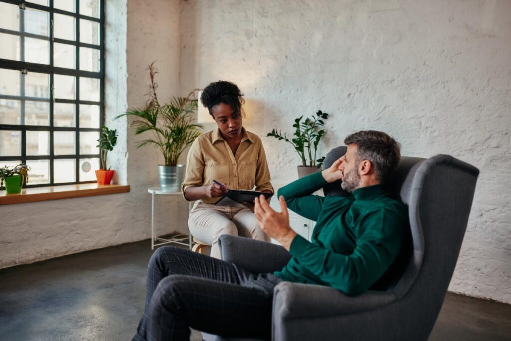 a specialist sits in a chair while a man sits in a chair in front of her as she discuss the difference between coaching and therapy for the man