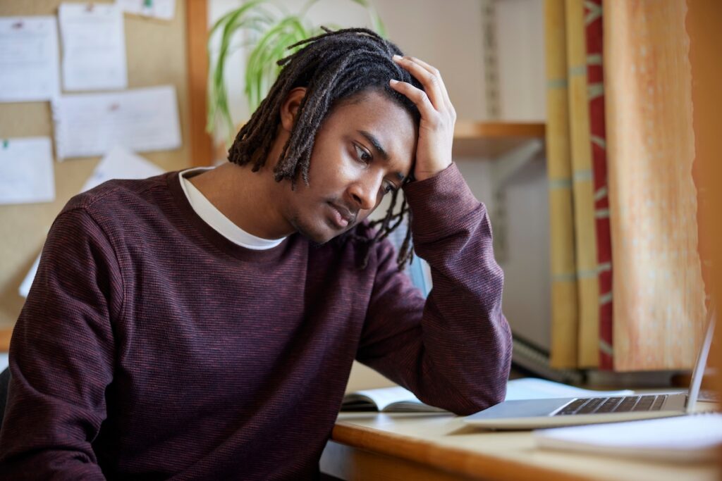 Young man sitting at a desk with a laptop rests his head on his hand, looking stressed or frustrated.